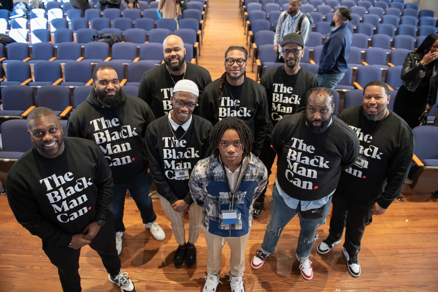 Black male educators in The Black Man Can shirts gathered around a young student at the 2026 Summit