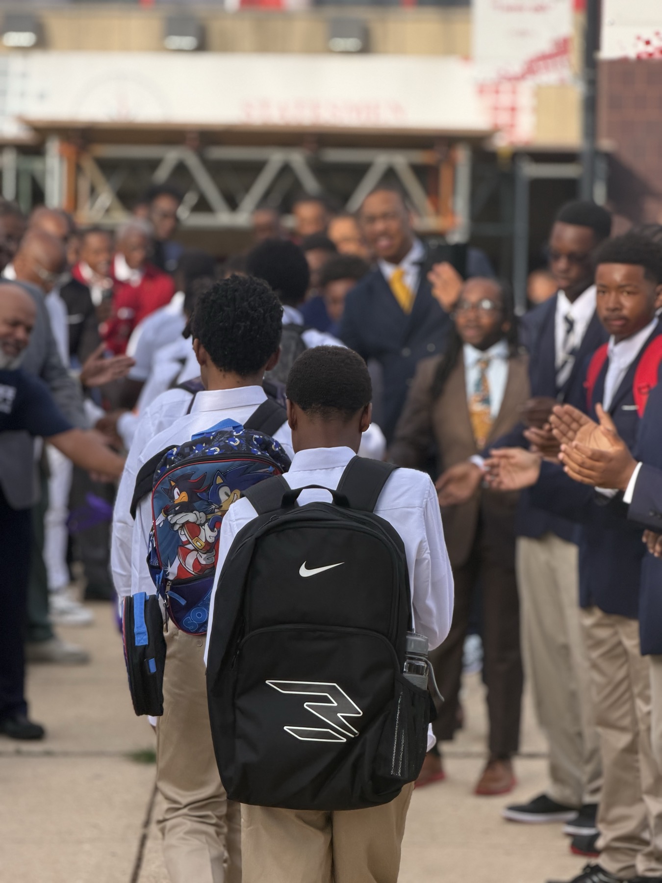 A boy with a backpack walks past men welcoming students to school